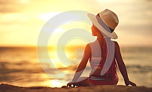 Happy child girl in swimsuit and hat sitting on beach at sunset