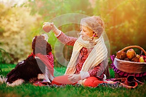 Happy child girl training her dog and giving him apple in sunny autumn garden