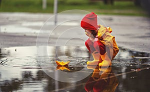 Happy child girl with umbrella and paper boat in puddle in a