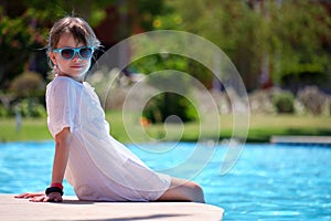 Happy child girl in white dress relaxing on swimming pool side on sunny summer day