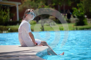 Happy child girl in white dress relaxing on swimming pool side on sunny summer day