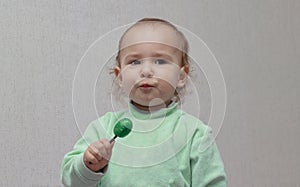 Happy child in a green jumper eating a green candy lollipop, on a light background. Beautiful little girl with brown eyes with a