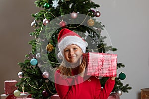 Happy child holding a box with a gift on the background of a Christmas tree. Little girl looking at the camera