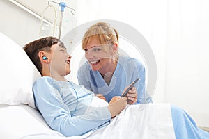 Happy child lying in bed in hospital room and smiling nurse using smartphone surfs the internet wearing earphones