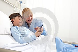 Happy child lying in bed in hospital room and smiling nurse using smartphone surfs the internet wearing earphones