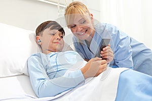 Happy child lying in bed in hospital room and smiling nurse using smartphone surfs the internet wearing earphones photo