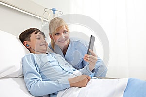 Happy child lying in bed in hospital room and smiling nurse using smartphone surfs the internet wearing earphones photo