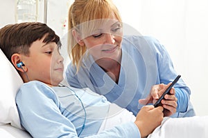 Happy child lying in bed in hospital room and smiling nurse using smartphone surfs the internet wearing earphones photo