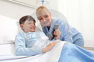 Happy child lying in bed in hospital room and smiling nurse using smartphone surfs the internet wearing earphones photo