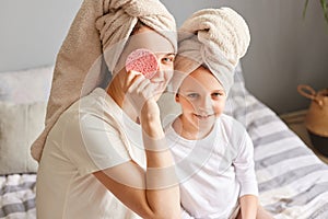 Happy child with mother sitting on bed in towels after morning shower having fun woman covering eye with cosmetic sponge family