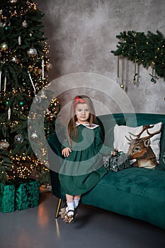 Happy child opens Xmas present.  Smiling little girl in dress sit with gift on sofa by Christmas tree with glowing garland lights.