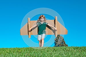 Happy child with paper wings against blue sky. Kid with toy jetpack having fun in spring green field outdoor. Freedom