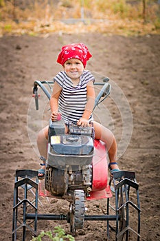 Happy child in red bandana and stripe tee sits on tiller on the field. baby girl driver cultivator