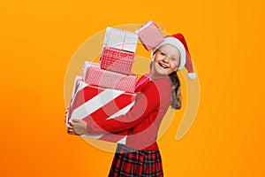 Happy child in red Santa hat holding a stack of Christmas presents. Little girl in the studio isolated on a yellow background
