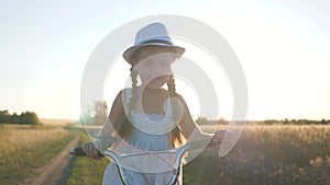 Happy child rides a bicycle in the park. A girl in a dress pedals in the sun. The kid is enjoying the dream. Cheerful