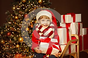 Happy Child in Santa Hat holding Gift. Stack of Xmas Presents on Christmas tree Lights Background