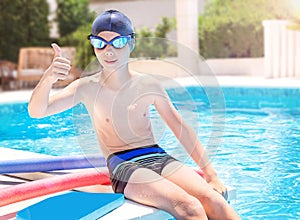 Happy child in sport goggles and cap showing thumbs up on the swimming pool background.