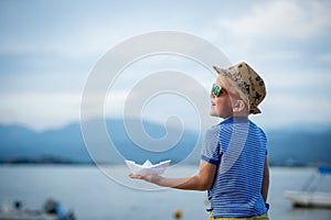 Happy child in sunglasses on beach. Summer vacation concept. Kid with paper boat in hand. Beautiful nature