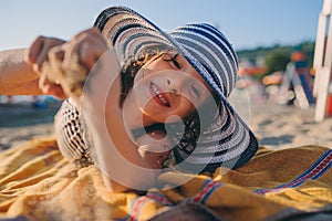 Happy child in swimsuit relaxing on the summer beach, lying on towel and playing with sand. Warm weather, cozy mood. Traveling on