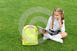 Happy child in uniform with school bag reading book sitting on green grass