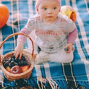 Happy childhood. A cute baby girl in a pink beanie sits on a blue blanket with a basket full of fruits