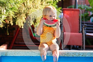 Happy childhood. Cute Caucasian little girl in yellow swimwear sitting by a pool and funny eating a slice of fresh