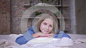 Happy childhood, portrait of beautiful little girl with blue eyes lying on pillow on bed in room