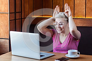 Happy childish young girl with blonde short hair in pink shirt is sitting in cafe and making video call on laptop, talking and