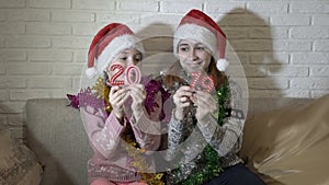 Happy children-girls show red figures 2019 to the camera. They sit in Santa`s hats and Christmas garlands on the couch