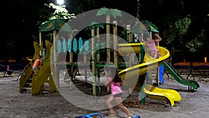 Happy  children have fun on slide at playground area at night. Three girls and a boy ride on a hill in the children`s town