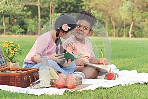 Happy children have a picnic in summer park, cute curly hair African girl with Asian buddy friend studying, reading book together