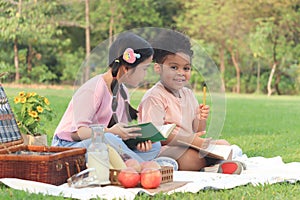 Happy children have a picnic in summer park, cute curly hair African girl with Asian buddy friend studying, reading book together