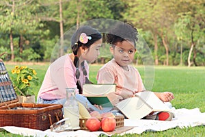 Happy children have a picnic in summer park, cute curly hair African girl with Asian buddy friend studying, reading book together