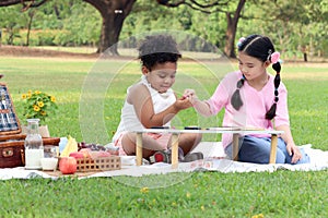 Happy children having a picnic in summer park, cute curly hair African girl with Asian buddy friend studying while sitting on mat