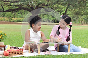 Happy children having a picnic in summer park, cute curly hair African girl with Asian buddy friend studying while sitting on mat