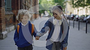 Happy childrens with backpacks back to home after study in elementary school holding hands. Caucasian boy and girl walks