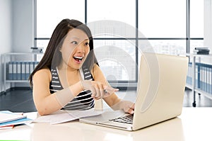 Happy chinese asian woman working and studying on her computer sitting at modern office desk smiling cheerful