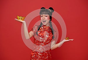 Happy Chinese new year. children asian girl wearing traditional qipao dress holding gold ingot isolated on red background