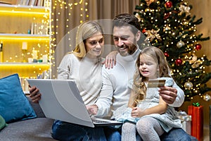Happy Christmas family doing online shopping sitting at home on sofa, couple husband wife and daughter with laptop and