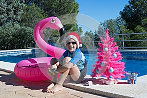 Happy Christmas Holidays. A cute child wearing a Santa hat by the swimming pool with a pink xmas tree and a flamingo buoy.