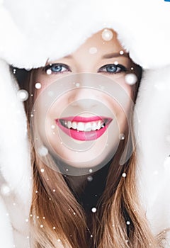 Happy Christmas and winter holiday portrait of young woman in white hooded fur coat, snow on blue background, fashion