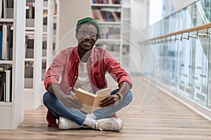 Happy clever black student man with book sitting on floor in university library, looking at camera