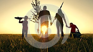 Happy, close-knit family with children, silhouette of farmers outdoors holding hands at sunset. Dad holds a young tree