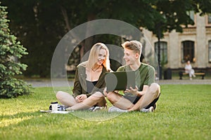 Happy college students communicating, talking, using laptop on campus lawn. Young cheerful caucasian couple of students watching