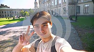 Happy confident asian guy is holding selfie camera and taking video in sunlights near beautiful old building, chilling