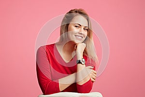 Happy confident blonde attractive woman keeps hand under chin, has shining smile, wears red sweater, isolated over pink background photo