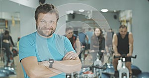 Happy confident coach posing at the background of blurred sportive people riding exercise bikes in gym. Smiling