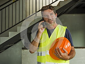 Happy and confident construction worker or solvent contractor man holding hardhat talking on mobile phone smiling cheerful working