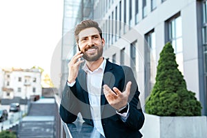 Happy confident european millennial bearded businessman in suit speaks by phone and gesticulates