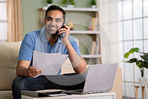 Happy confident indian man talking on mobile phone by holding insurance or property documents at home - concept of